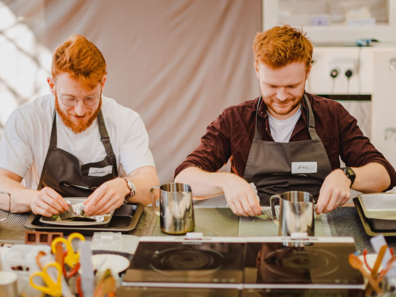 Two men are sat at a table wearing aprons with candle making equipment